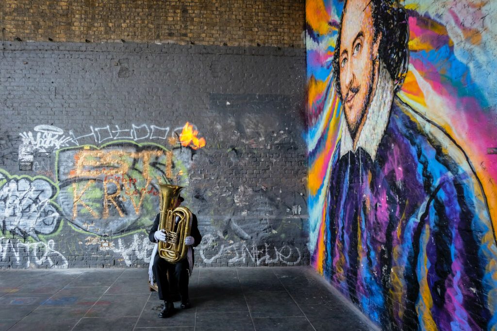 Tuba player near a shakespeare mural surrounded by grafitti